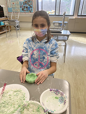 A little girl with a tie dye sweatshirt that has a peace sign on it is wearing a mask and holding a small green bowl.