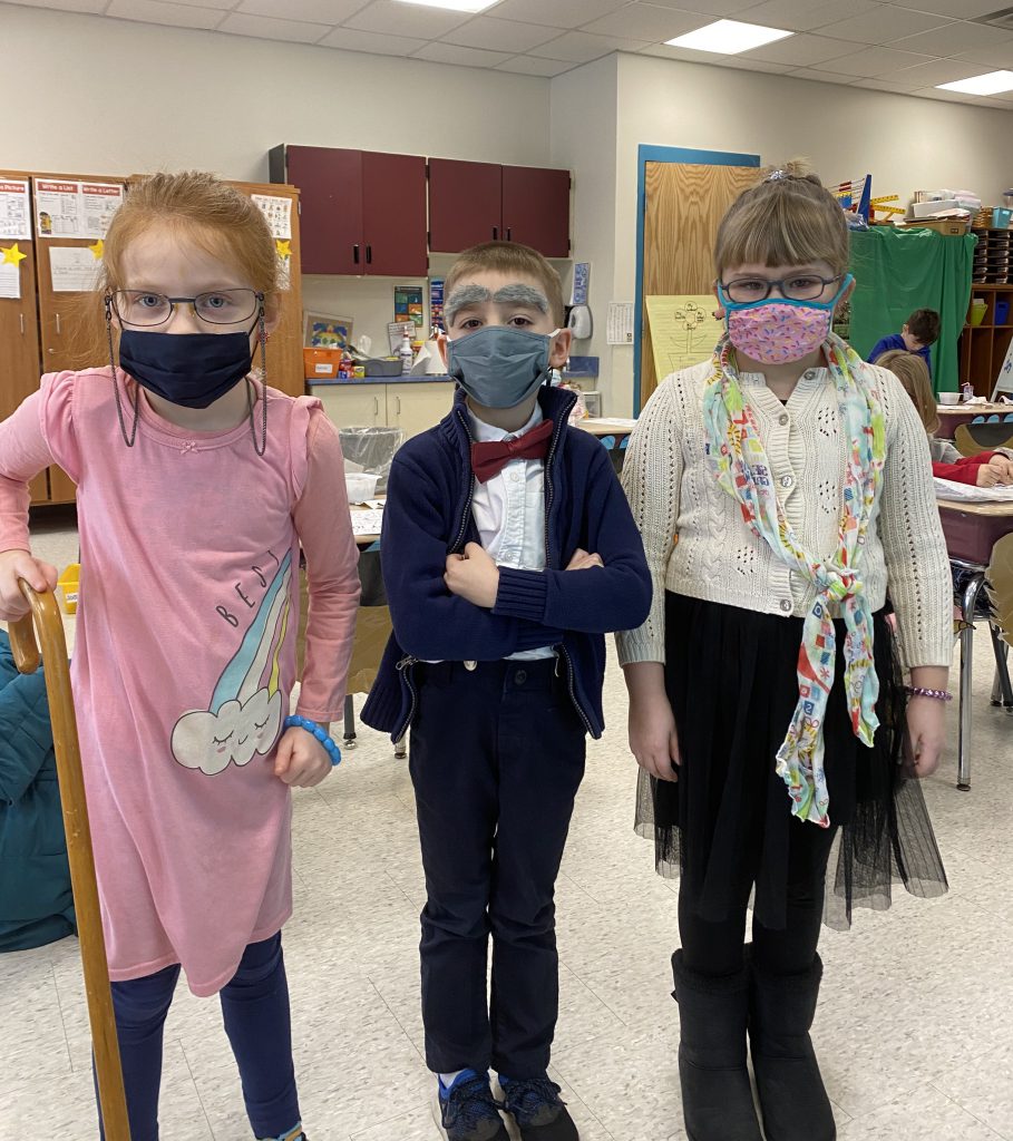 Three elementary age children dressed as old people. One has a cane, another has bushy gray eyebrows and wears a bow tie.