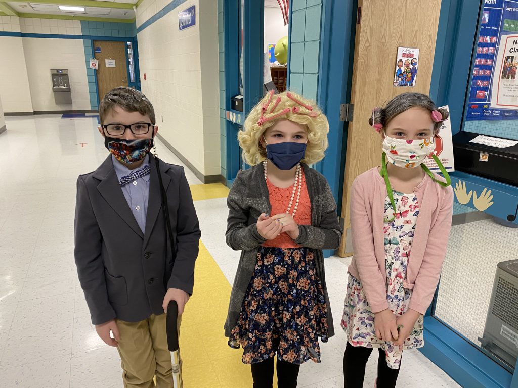 Three elementary school kids dressed as 100 year old people. Boy has a cane, bow tie and jacket. Two girls have wigs with curlers in their hair, wearing sweaters and pearls.