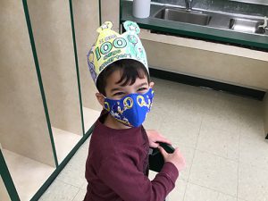 A boy wearing a maroon long-sleeve shirt, blue mask and a hand-colored crown that says I'm 100 days smarter.