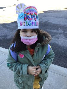 A little girl with dark hair, wearing a jacket and pink mask and a hand-colored crown that says I'm 100 days brighter.