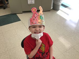 A boy wearing a red shirt and a hand-colored crown that says I'm 100 days smarter. He is tugging on his mask and smiling broadly.
