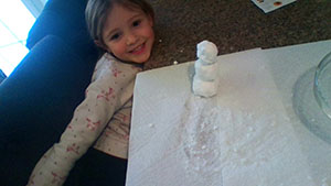 A girl with long brown hair smiles. She has a little snowman on the table in front of her.