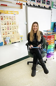 A woman with long blonde hair sits in an office chair smiling. She is wearing dark clothes and there is a whiteboard next to her with various photos on it.