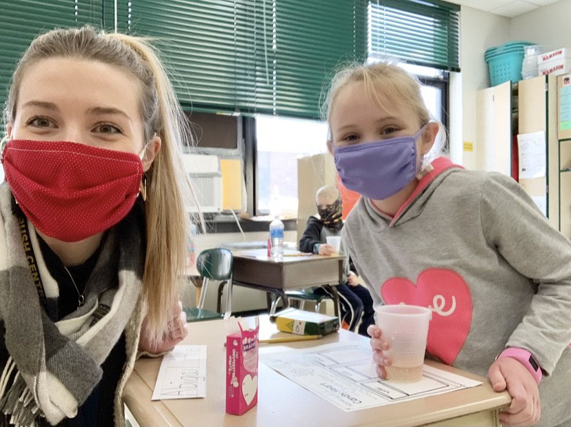 A woman with long blonde hair wearing a red mask is with a student also with blonde hair wearing a purple mask, gray sweatshirt with a pink heart on it.