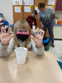 A blond girl holds up her hands with lots of snow-like material on them. She is wearing a black and pink mask.