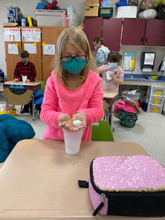 A blonde girl wearing a bright pink shirt and a green mask holds a tiny snowball in her hands.