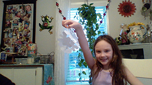 A girl with long brown hair wearing a pink shirt holds up a paper snowflake.