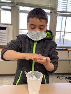 A boy wearing a mask and a gray sweatshirt rolls his homemade snow in his hands. There is a cup in front of him with more of the snow in it.