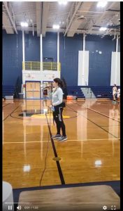 A woman wearing black athletic pants and gray sweatshirt holds a microphone and sings in a gymnasium.