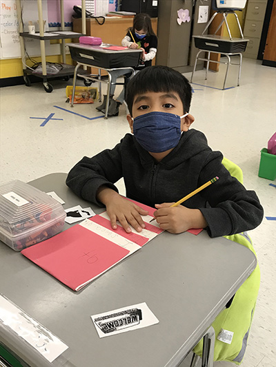 A boy with a dark shirt and mask holds a pencil and writes on a red and white paper hat.