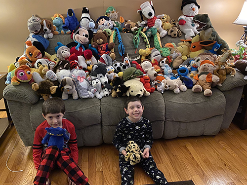 Two little boys sit on the floor with a couch behind them with 100 stuffed animals covering it.