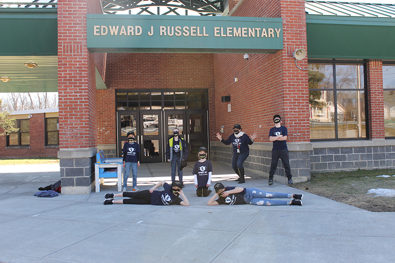 Six elementary students and one adult are outside of a brick building with the name Edward J. Russell elementary above. Two of the students are lying down in front. All are wearing black shirts.
