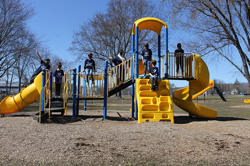 Six elementary students and one adult are in different spots on a playground jungle gym. Blue sky is above them and a large tree behind them.