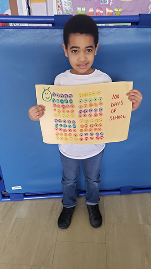 A boy wearing a white shirt and blue jeans holds up a large piece of cardboard with rows of colorful cereal on it totaling 100