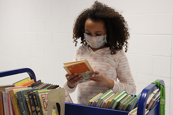 A girl with dark curly shoulder length hair, wearing a multi-colored mask and a light colored shirt, holds a book she took from a cart.