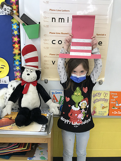 A girl dressed in a shirt with the grinch on it, holds up a red and white striped paper hat over her head. She is wearing a mask.