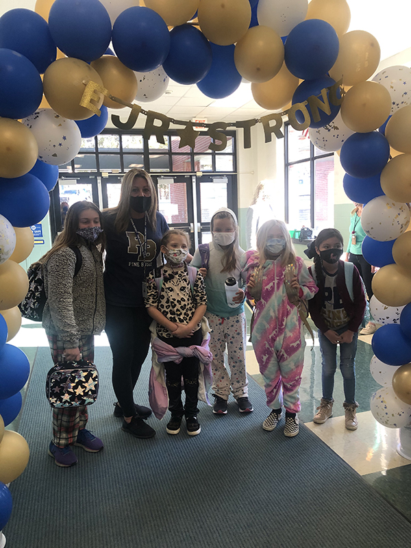 A group of five students and one adult under a blue and gold balloon arch. All are wearing masks and dressed in pajamas.