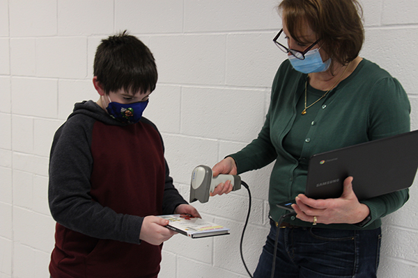 A woman wearing glasses, mask and a green sweater holds a scanner as she scans a book being held by a boy wearing a dark sweatshirt and mask.
