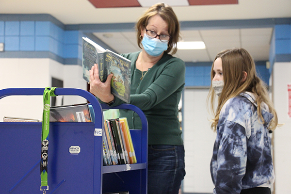 A woman wearing a green sweater, mask, glasses and with short blonde hair holds a book open above a cart with many other books on it. A student with long blonde hair, mask and a blue sweatshirt looks at the open book.