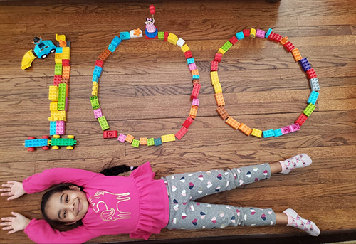 A smiling girl wearing a pink shirt is lying down on a wood floor. Just above her on the floor are 100 legos in the shape of the number 100.