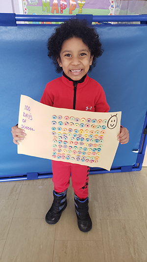 A little boy dressed in red is smiling broadly as he holds a large piece of cardboard with 100 colorful pieces of cereal .