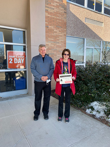 A high school age woman in a red coat holds a certificate and stands next to a man wearing a blue jacket and black pants. They are standing in front of a building.