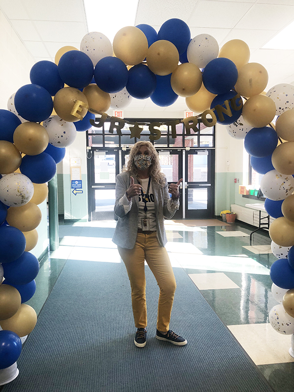 A woman with long blonde hair, yellow pants and wearing a mask is under an arch of blue and gold balloons giving thumbs up.