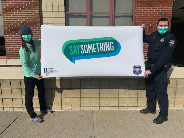 A woman at left wearing green shirt and mask and a man at right in a police officer's uniform, hold a white banner with the words say something in the center.