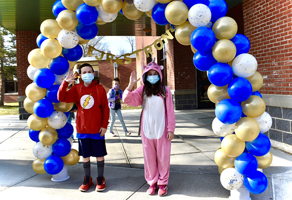 A boy and girl stand under a blue and gold balloon arch. The girl is in pink and white pajamas with a pink hood. The boy has a red shirt with a gold lightning bolt on it and shorts. Both are wearing a mask. The picture was taken outside with a brick building in the background.