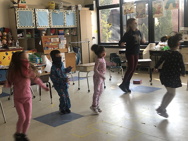 A group of kindergarten students dressed in their pajamas, all jumping up and down to an exercise class. Their teacher, a woman dressed in blue, is also jumping.