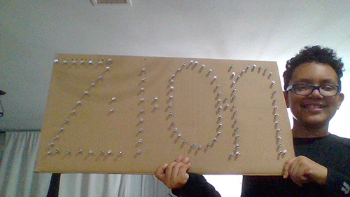 A fifth grade boy wearing glasses smiles and holds up a piece of cardboard with his name, Zion, spelled out in pushpins.