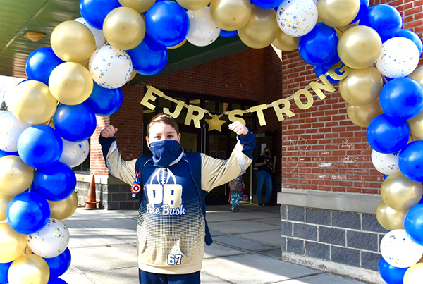 A boy in a gold sweatshirt that says PB holds up his arms as if flexing his muscles. He is wearing a blue mask and under an arch of blue and gold balloons with the words EJR Strong on it.