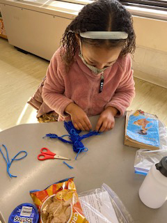 A girl in a pink jacket, white headband, and a mask sits at a table and makes a yard doll out of blue yarn. There are scissors and a copy of a book on the table.