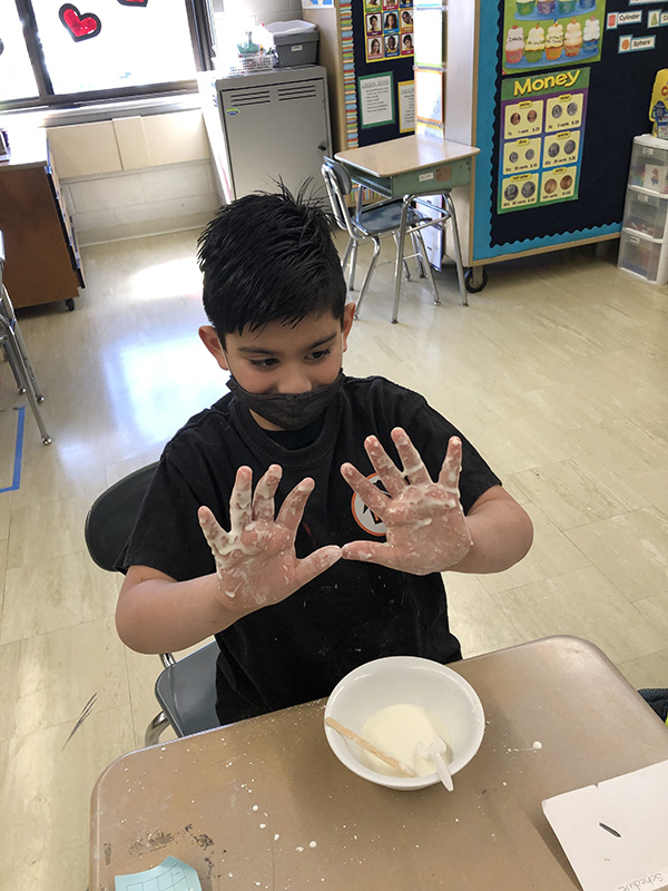 A boy with black mask and t-shirt holds up his hands with gooey stuff on it. A bowl sits on his desk with the off-white sticky substance.