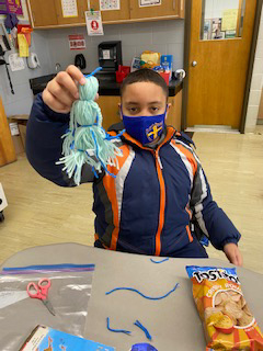 A boy wearing a blue jacket and a blue mask holds up a light blue yard doll he just made. On the table are snacks and scissors.
