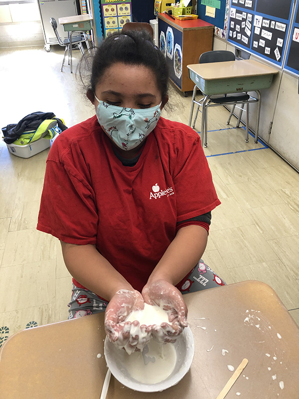 A girl with her dark hair pulled back, wearing a red t-shirt and blue mask holds her two hands together with a pool of off-white liquid in it.