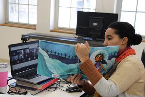 A woman wearing a tan sweater and red scarf holds up a large book to screens with her students watching from home.