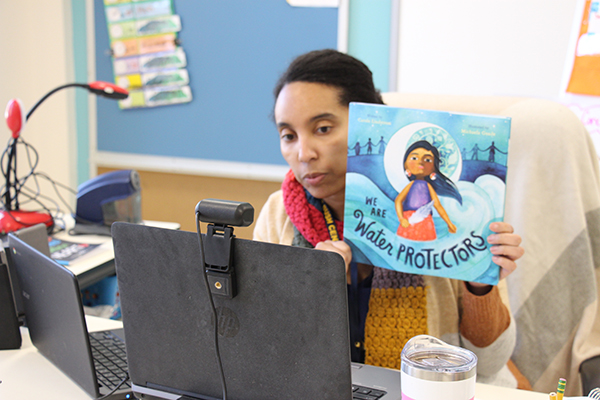 A woman holds up a book called We are Water Protectors. It is different shades of blue with a young girl dressed in native American clothing in the center.