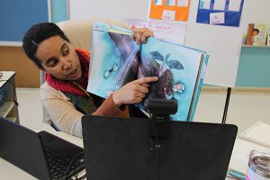 A woman wearing tan sweater and red scarf holds a book up and points to something on the open page as her students watch remotely. The computer screens are in front of her.