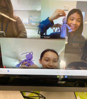 A computer screen shows several students smiling and holding up purple yarn dolls.
