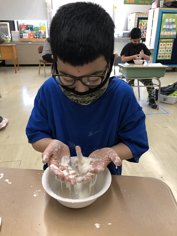 A boy with a blue shirt, glasses and a mask holds his hands over a white bowl that is filled with an off-white material. The liquid is on his fingers and dripping into the bowl.