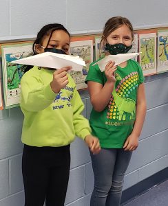 Two second graders, both dressed in green shirts and wearing masks, hold up their paper airplanes. 