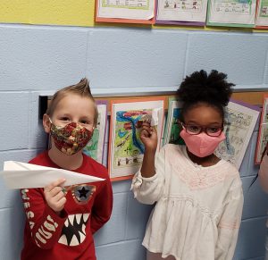 Two second grade students, a boy in a red shirt and a girl with glasses and a white shirt, hold up their paper airplanes. Both are wearing masks.