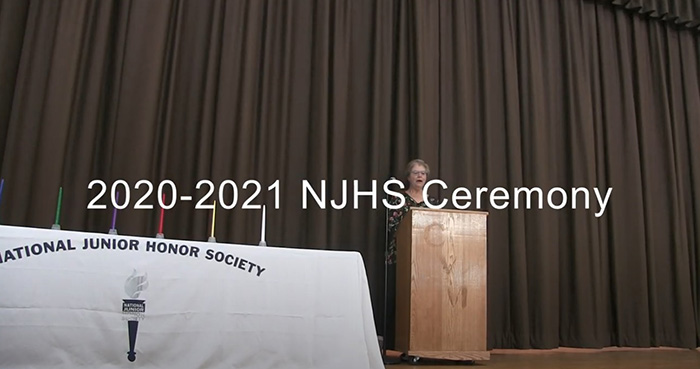 A woman stands at a podium with a table on the left. It has a white table cloth with the words National Junior Honor Society on the front and five long, large candles on top.