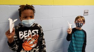 Two young boys hold up their paper airplanes. One is wearing a black shirt with a graphic on it and the other has blue and green stripes. Both are wearing masks.