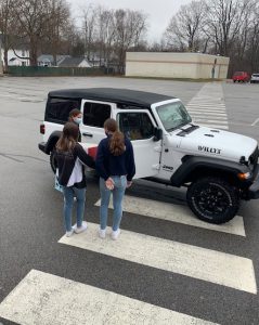Two students, one holding a large box, approach a white jeep with a black roof.