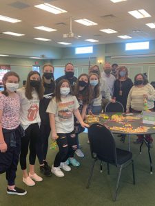 A group of 11 people, all with masks on, stand around a table with candy and bags on it.