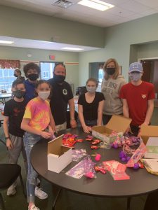 Seven people, kids and adults stand around a table that has candy and Easter bags on it. They are all wearing masks.