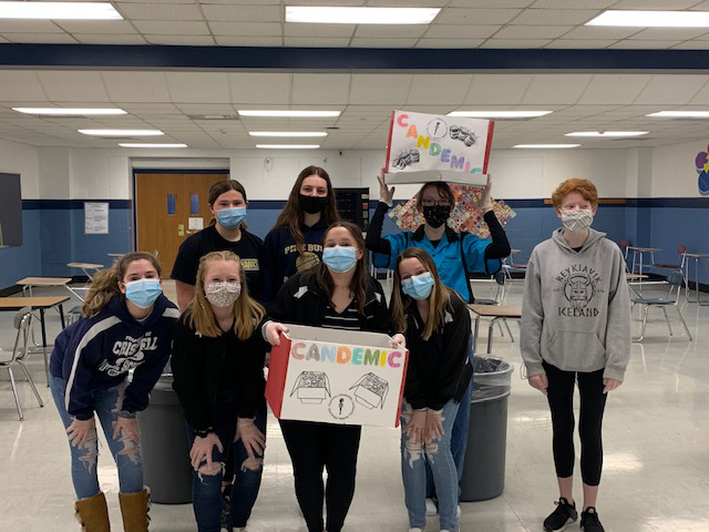 Eight middle school students stand together, each wearing a mask. One holds up a sign and another a large box that say Candemic on them.